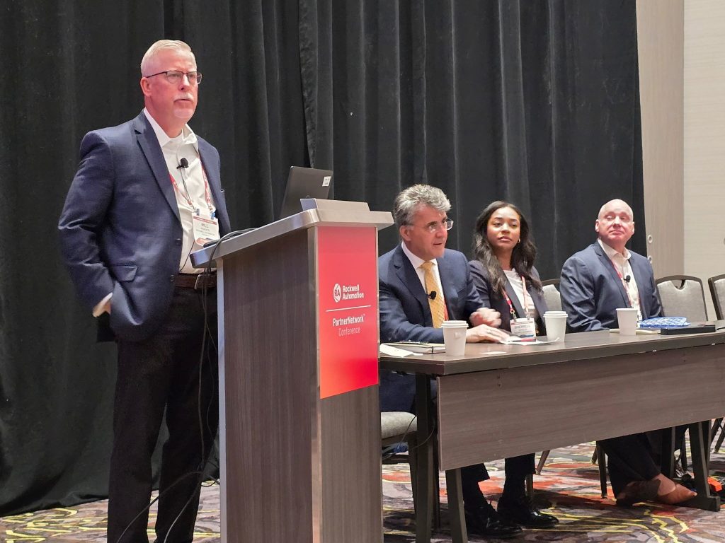 Speaker presenting at a conference podium beside a seated panel